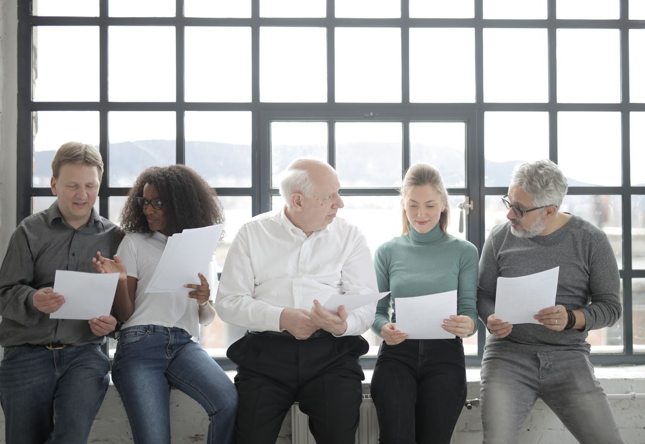 Cheerful multiracial coworkers in casual wear gathering together and sitting with papers near big fenced window in sunlight and looking at each other